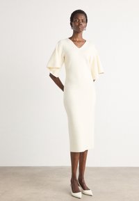 Fitted cream dress with a V-neck, short bell sleeves, knee-length hem, and smooth texture. Worn with pointed white heels.