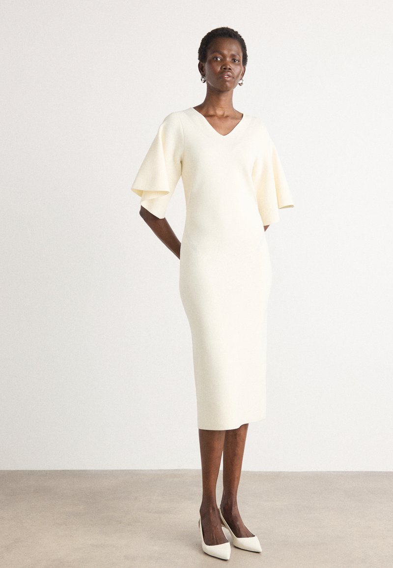 Fitted cream dress with a V-neck, short bell sleeves, knee-length hem, and smooth texture. Worn with pointed white heels.