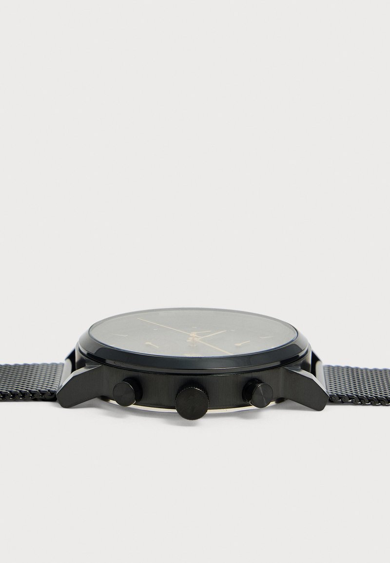 Vue latérale d'une montre-bracelet noire avec un bracelet en maille métallique et trois boutons sur le boîtier sur fond blanc.
