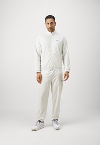 Fato de treino cinza claro feito de tecido suave com um padrão de grelha subtil. Apresenta um fecho frontal, punhos elásticos e um logo bordado no peito.