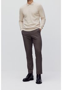 Homme portant un polo beige à manches longues, un pantalon marron coupé sur mesure et des bottes en cuir noir à lacets, debout les mains dans les poches.