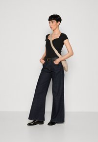 Body noir à côtes, jeans larges bleu marine, chaussures noires brillantes et sac en bandoulière beige. Pose debout contre un fond uni.