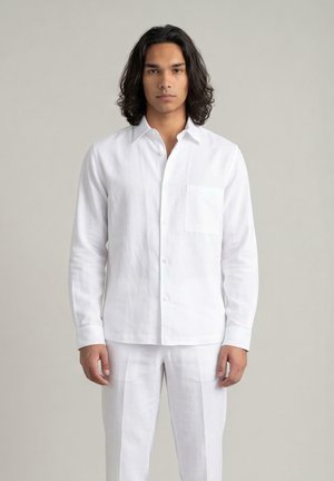 Man met donker schouderlang haar die rechtop staat, gekleed in een wit overhemd met lange mouwen en witte broek tegen een effen achtergrond.