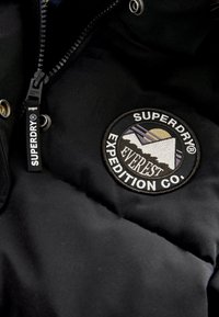 Veste noire avec fermeture éclair Superdry et patch circulaire Everest Expedition Co. montrant une broderie de montagne et de soleil.