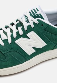 Grön mockaskor med vita snören och en framträdande vit "N"-logotyp. Har vadderad tunga och texturerad gummisula.