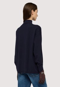 Maglione di maglia oversize blu navy con colletto alto, spalle basse e finitura texturizzata. Il modello indossa guanti di pelle marroni e jeans azzurri.