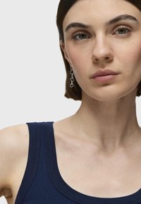 Top de tirantes azul marino con escote en forma de scoop. La modelo lleva pendientes colgantes de plata con acentos en forma de nube enlazados. Textura de piel suave.