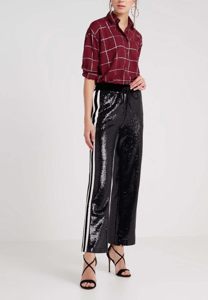 Chemise à carreaux rouges avec manches retroussées ; pantalon noir à sequins avec bandes blanches latérales ; sandales à talons hauts noires à brides.