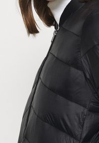 Svart vadderad jacka med dragkedja framtill, med quiltade sektioner och hög krage. Slät textur med glänsande finish.