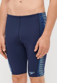 Shorts de bain navy avec une coupe ajustée. Fabriqués en matériau extensible. Présentent des motifs en zigzag bleus sur les côtés et un logo sur la jambe inférieure.