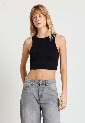 Giovane donna con indosso un top corto nero senza maniche e jeans grigi a vita alta, in piedi con un'espressione neutra davanti a uno sfondo semplice.