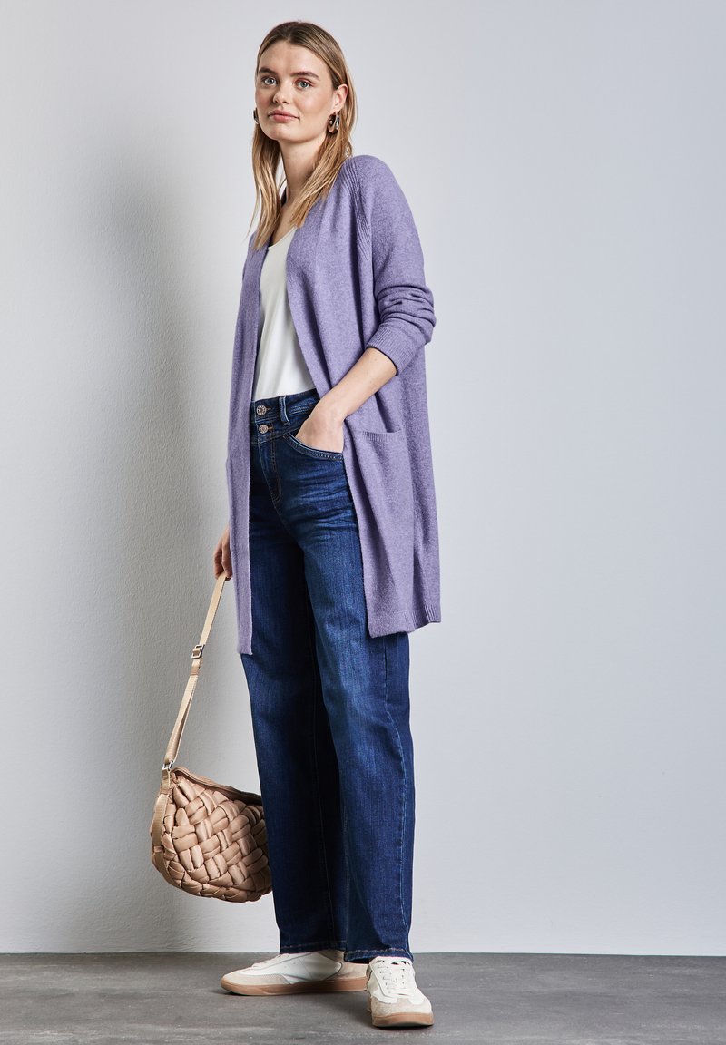 Lavendel lång kardigan, vit topp, mörkblå jeans och vita sneakers. Vävd beige väska med lång rem. Släta texturer, avslappnad passform.