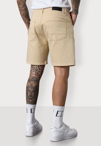 A person stands wearing beige shorts and white sneakers. Their leg features intricate tattoos, and they are barefoot on a neutral background.
