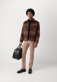 Bruin en rood geblokt wollen jack, zwarte coltrui, beige broek, zwarte chelsea boots en een zwarte leren tas. Eenvoudige ontwerp elementen zichtbaar.