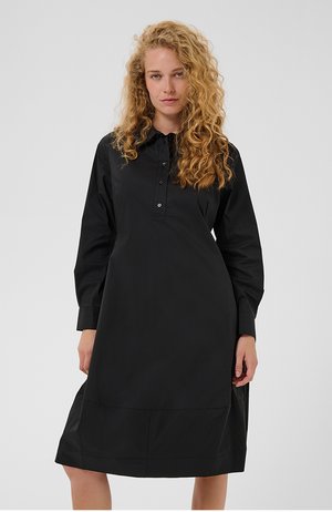 Kobieta o kręconych blond włosach, ubrana w czarną sukienkę koszulową z długim rękawem, stojąca na tle jasnego, jednolitego tła.