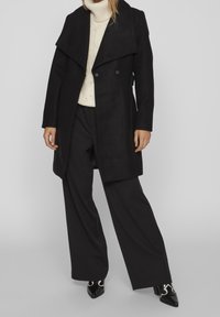 Cappotto nero doppiopetto con colletto alto, abbinato a un maglione crema a trecce e pantaloni neri a gamba larga. Scarpe nere a punta.