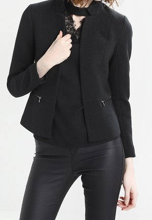 Frau trägt einen eng anliegenden schwarzen Blazer über einem schwarzen Spitzen-Top und schwarzen Lederhosen, posiert mit einer Hand nahe dem Kragen.