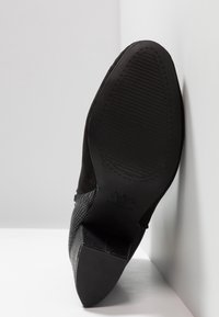 Chaussure noire à talon avec semelle texturée et bout pointu, positionnée à la verticale contre un mur d’angle blanc et un sol.