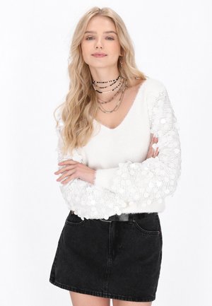 Pulover alb tricotat cu decolteu în V și mâneci texturate, având motive circulare din dantelă. Asortat cu o fustă neagră din denim.