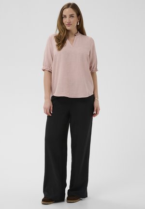 Mujer de pie con blusa de manga corta rosa claro, pantalones anchos negros y zapatos marrones frente a un fondo gris liso.