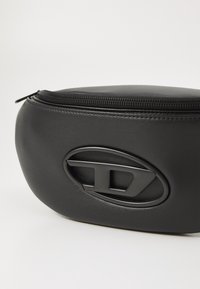 Schwarze, oval geformte Tasche aus synthetischem Material, mit einem Reißverschlussverschluss und einem markanten schwarzen Logo-Element auf der Vorderseite.
