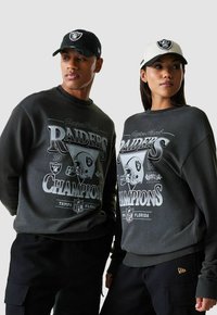 Felpe grigie con un grafiche dei Raiders e la scritta "Campioni", abbinate a pantaloni cargo neri. Cappi bianchi e neri completano il look.
