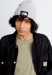 Beanie grigia con logo, giacca di jeans nera e t-shirt beige. Capelli ricci e piercing al setto nasale visibili. Sfondo semplice. Nessun dettaglio aggiuntivo.