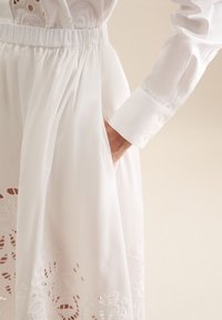Hvid kjole med elastisk talje, der har detaljeret broderi langs kanten og lange ærmer med knapdetaljer.
