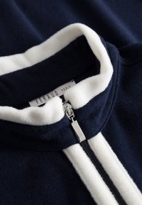 Navy blue fleece zip-up jacket with a white collar and front stripes, featuring a silver zipper and brand label inside.