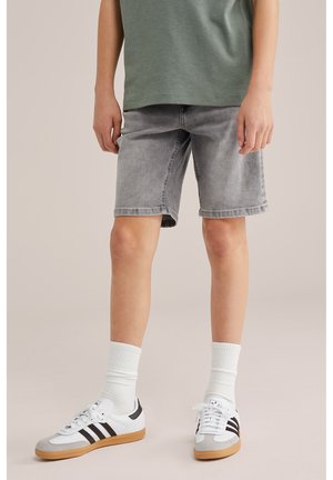 Niño con pantalones cortos de mezclilla grises, camiseta verde, calcetas blancas hasta media pierna y zapatillas blancas con rayas negras, de pie sobre un suelo beige.