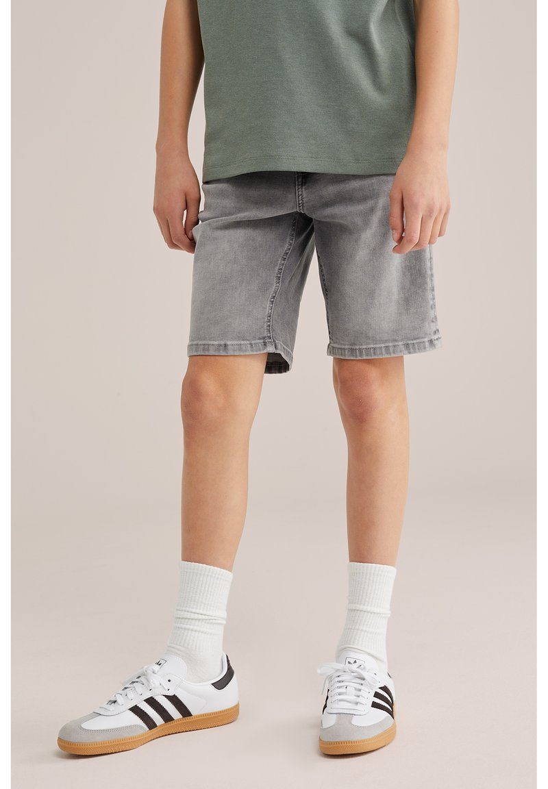 Kind draagt grijze spijkershorts, groen T-shirt, witte sokken, en witte sneakers met zwarte strepen, staand op een beige vloer.