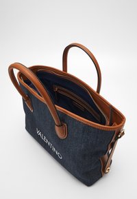 Denimtrendväska med bruna läderhandtag, stickade detaljer och innerfickor. Har en vit logotyp på framsidan. Öppen toppdesign.