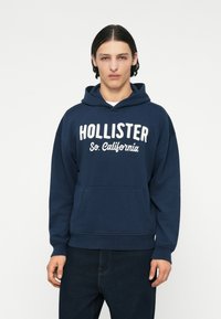 Mornarica kapuca iz bombaža, z sprednjo žepom in belim napisom "HOLLISTER So. California" čez prsi. Ribljen rob in zapestja.