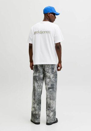 Mann trägt weißes Jack & Jones T-Shirt, graue Acid-Wash-Jeans, blaue Kappe, schwarze Sonnenbrille und schwarze Schuhe, steht mit dem Rücken zum Betrachter vor weißem Hintergrund.