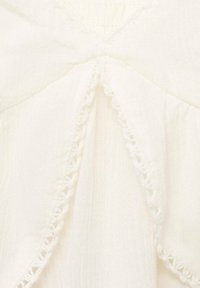 White top featuring a bow detail at the chest, with textured fabric and intricate lace trim along the edges. Soft and lightweight appearance.