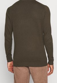 Pull vert olive à manches longues, poignets et ourlet côtelés, en maille lisse, porté avec un pantalon en velours côtelé beige texturé.