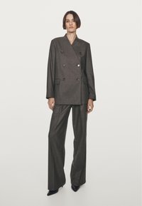 Blazer grigio sartoriale doppiopetto con bottoni, abbinato a pantaloni grigi a gamba larga e tacchi neri a punta. Tessuto morbido.