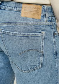 Jeans de denim azul claro con una etiqueta de cuero en la cintura con el texto "G-STAR ORIGINALS RAW DENIM" grabado. Bolsillos estándar y detalles de costura visibles.