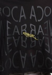 Černé zkrácené tričko s hladkou texturou, které má tučný, překrývající se text v tmavě šedé a žluté logo "ROC AN WEAR" ve středu.