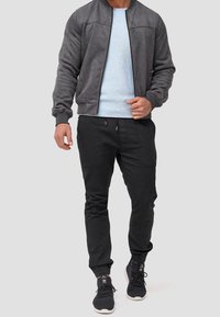 Veste bomber grise, pull bleu clair, pantalon noir, baskets noires. La veste possède des poignets côtelés et une fermeture éclair ; le pantalon a des poches et des poignets élastiques.