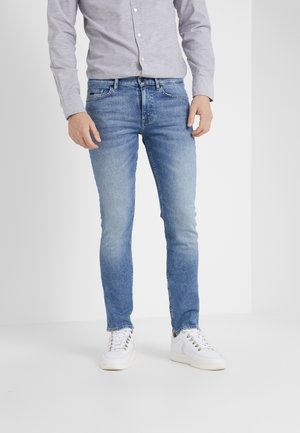 Mężczyzna ubrany w jasnobłękitne dżinsy slim fit, szarą koszulę zapinaną na guziki i białe trampki na jednolitym jasnym tle.