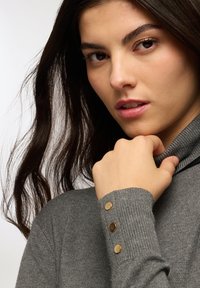 Maglione collo alto grigio realizzato in tessuto a costine, con tre bottoni dorati sul polsino. Texture morbida e design aderente.