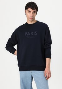 Felpa blu navy con polsini e orlo a costine, presenta la scritta "PARIS" ricamata in un blu chiaro a contrasto sul davanti.