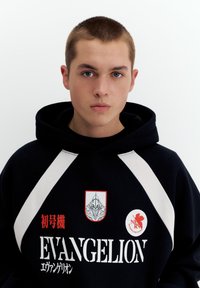Ung mand med kort hår iført en sort hættetrøje med hvide striber og røde og hvide Evangelion-tema logoer og tekst.