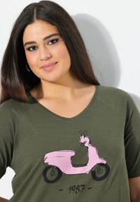 Camiseta verde oliva de cuello en V con un gráfico de un scooter rosa y el número "1967" en negro; tela suave y corte relajado, que acentúa el diseño.