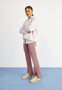 Světlofialová mikina s volným střihem, přehozená přes proužkovanou košili na knoflíčky. Růžové rozšířené kalhoty s postranními rozparky, doplněné béžovými teniskami.