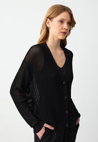 Cardigan nero lavorato a maglia con un design testurizzato e a trama aperta, dotato di scollo a V e sei bottoni frontali. Le maniche lunghe si stringono al polso.