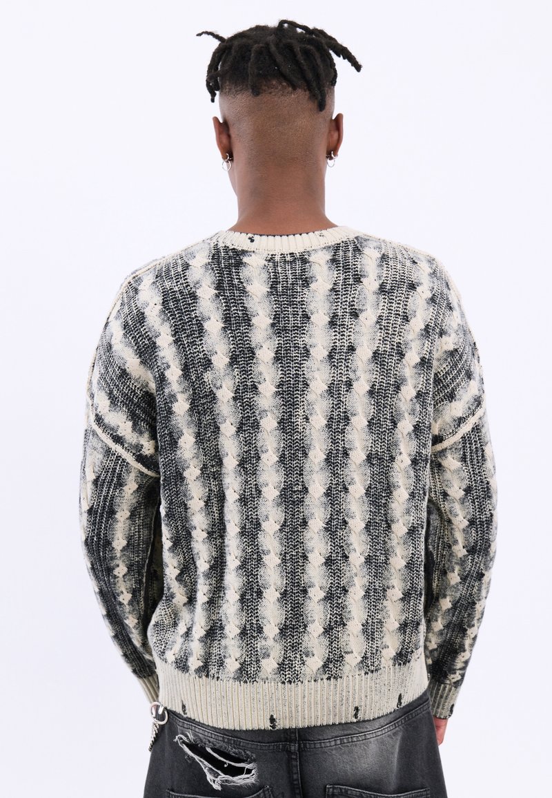 Personne aux cheveux courts torsadés portant un pull en maille rayé noir et blanc et un jean noir usé, de dos sur un fond blanc.
