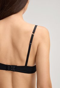 De rug van een vrouw die een verstelbare zwarte bh-band en haak-en-oogsluiting toont tegen een lichte effen achtergrond.