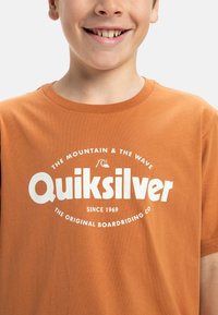 Krátky rukáv oranžové tričko vyrobené z bavlny, s bielym textom loga a kruhovou grafikou. Hladká textúra, okrúhly výstrih.